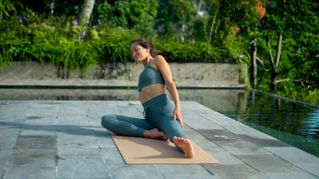 A young attractive European brunette sports girl makes yoga in nature with a green rainforest on a lit background by the pool on a mat and breathes deeply.の写真素材
