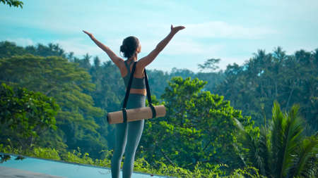 A young attractive European brunette sports girl goes with a yoga rug in nature with a green rainforest on a lit background by the pool.の写真素材