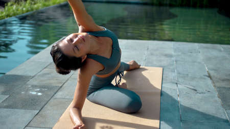 A young attractive European brunette sports girl makes yoga in nature with a green rainforest on a lit background by the pool on a mat and breathes deeply.の写真素材