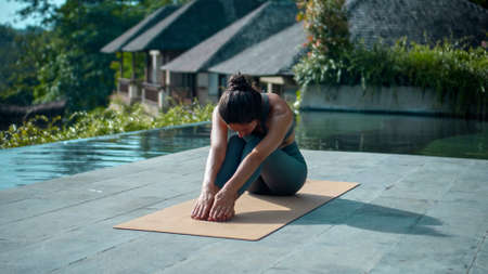 A young attractive European brunette sports girl makes yoga in nature with a green rainforest on a lit background by the pool on a mat and breathes deeply.の写真素材