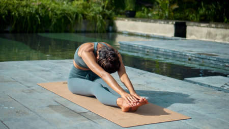 A young attractive European brunette sports girl makes yoga in nature with a green rainforest on a lit background by the pool on a mat and breathes deeply.の写真素材