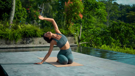 A young attractive European brunette sports girl makes yoga in nature with a green rainforest on a lit background by the pool on a mat and breathes deeply.の写真素材