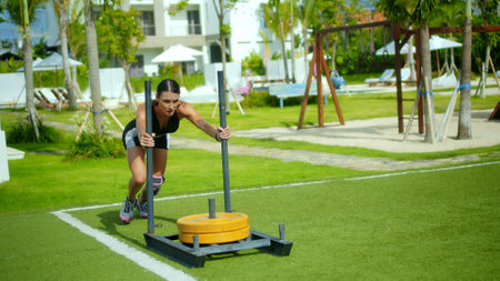 Young sports European brunette girl pushes steel sleigh for crossfit on green lawn with weightsの写真素材