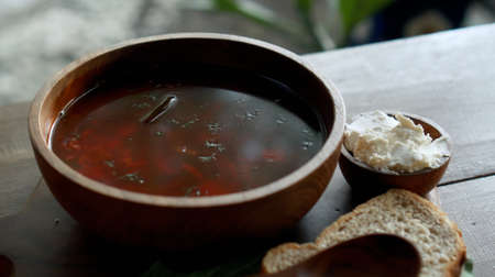 Traditional Russian fresh hot soup borsch with beetroot, red color, with sour cream and fresh bread in a wooden plate standing on a wooden table in the villageの写真素材