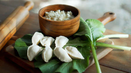 Traditional rustic Russian fresh handmade dumplings lying on a wooden table on a cutting board with cottage cheese in a wooden plate and spinachの写真素材