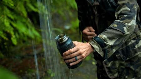 Hands of young hiker hold glass, draws water from waterfall for drinking, in the middle of...