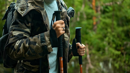 Hands of European young hiker in Hakki-colored robe hold rainforest walking sticks with green plants in backgroundの写真素材