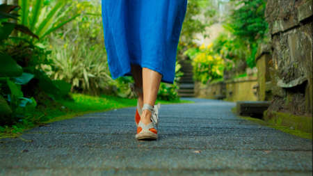 Legs of a European girl in a blue dress walking in shoes along the road with antique stonework along a green parkの写真素材