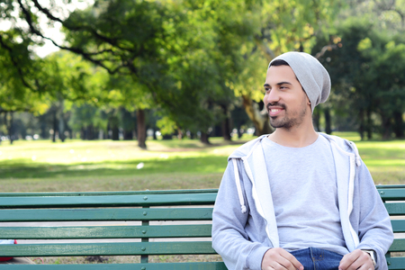 Portrait of young latin man sitting on park bench. Outdoors.の写真素材