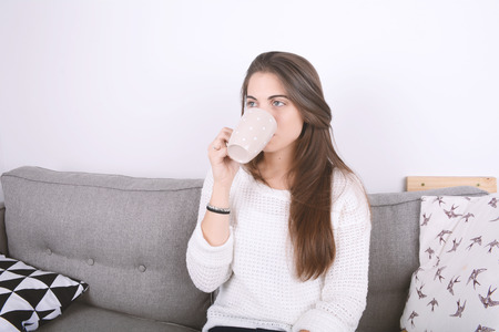Beautiful young woman drinking coffee and relaxed on couch. Indoors.の写真素材