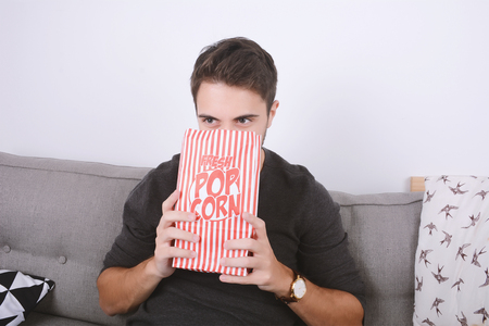 Young man eating popcorn and watching movies relaxed on couch. Indoors.の写真素材