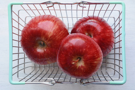 Shopping basket filled with Ripe red apples on white wooden background.