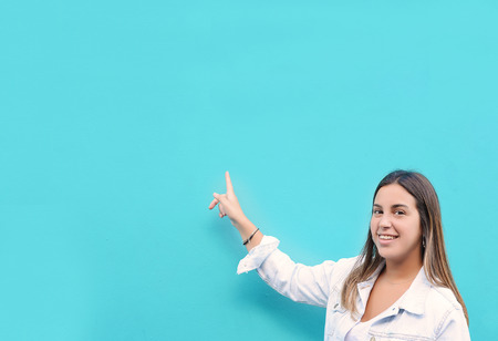 Portrait of smiling young woman pointing at copy space on light blue wall. Advertising conceptの写真素材