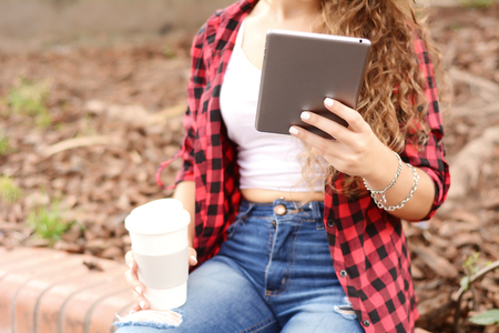 Young student girl with a tablet and a disposable coffee cup sitting on the park in autumn.の写真素材