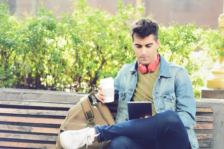 Young man sitting on a bench with tablet and drinking coffee in the park. Outdoorsの写真素材