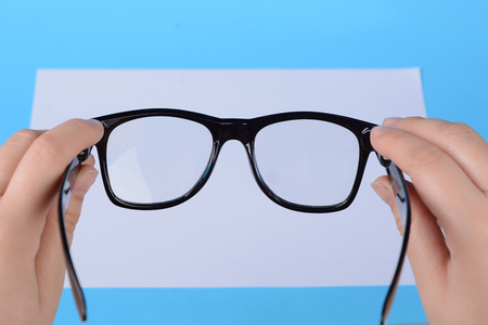 Eyeglasses in a female hand over a blank paper. Focus on eyeglasses. Light blue backgroundの写真素材