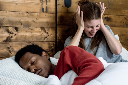 Portrait of irritated woman blocking ears with hands while man snoring on bed. Multiracial couple in bed at home.の写真素材