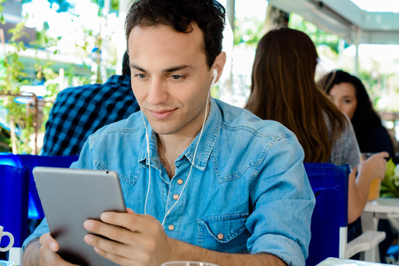Young latin man with earphones using digital tablet in coffee shop.