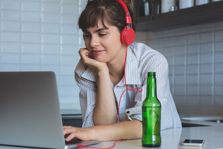 Young latn woman with beer and using her laptop pc in the kitchen at home.の写真素材