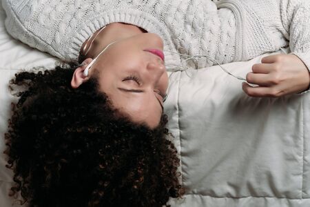 Young beautiful woman lying on bed and listening to music with headphones. Smiling woman enjoying...
