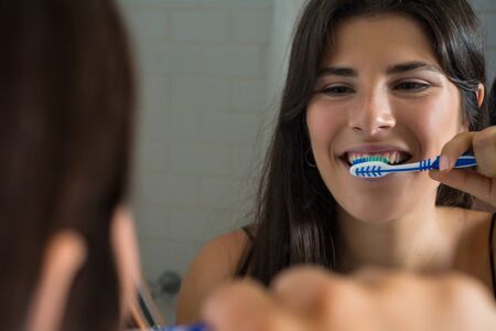 Smiling young woman with toothbrush brushing teeth in front of mirror. Health care and oral hygiene...
