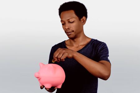 Portrait of young american man putting a coin in a piggy bank on studio. Save money concept.の写真素材