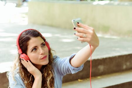 Young latin woman with red headpones making a selfie with mobile phone. Outdoorsの写真素材