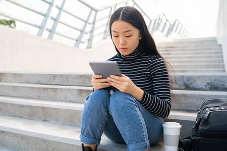 Portrait of young Asian woman using her digital tablet while sitting at stairs outdoors. Technology concept.の写真素材