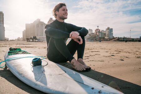 Young surfer sitting on sandy beach looking at the ocean and next to his surfboard. Sport and water sport concept.の写真素材