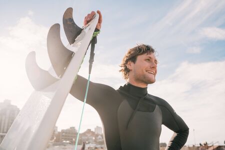 Close-up of young surfer standing at the beach with his surfboard and wearing black surfing suit. Sport and water sport concept.の写真素材