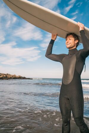 Portrait of young surfer at the beach holding up his surfboard and wearing a black surfing suit. Sport and water sport concept.の写真素材