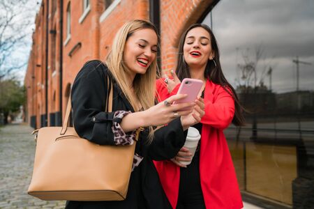 Portrait of two young friends using their mobile phone outdoors at the street. Lifestyle and friendship concepts.の写真素材