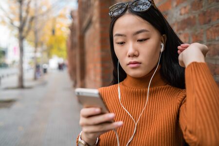 Portrait of young Asian woman using her mobile phone with earphones outdoors in the street. Urban and communication concept.の写真素材