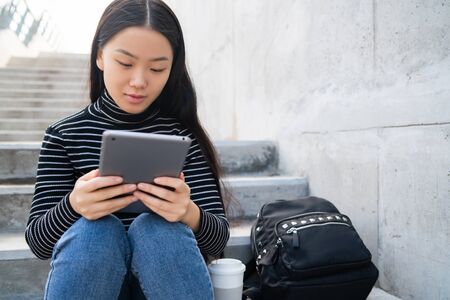 Portrait of young Asian woman using her digital tablet while sitting at stairs outdoors. Technology...