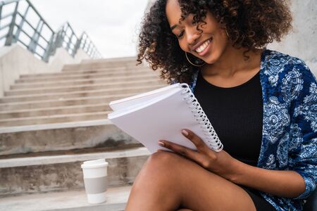 Portrait of young afro-american woman writing in a notebook while sitting outdoors on stairs.の写真素材