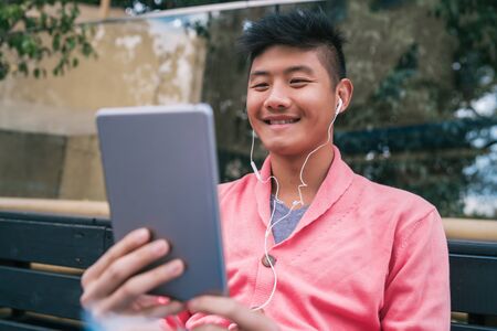 Portrait of young Asian man using his digital tablet with earphones while sitting in a bench outdoors. Technology concept.の写真素材