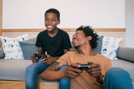 Portrait of happy African American father and son sitting in sofa couch and playing console video...