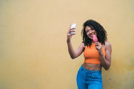 Portrait of afro woman taking selfies with her mophile phone while eating ice-cream. Technology and...