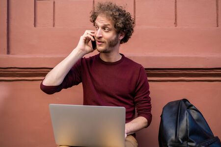 Portrait of young man talking on the phone and using his laptop while sitting outdoors. Technology...