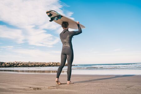 Portrait of young surfer at the beach holding up his surfboard and wearing a black surfing suit....