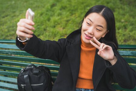 Portrait of young beautiful Asian woman taking a selfie with her mobile phone while sitting on a...