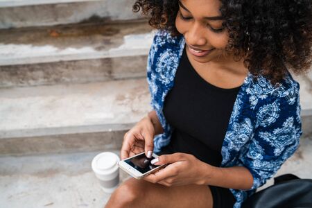Portrait of young beautiful afro american woman using her mobile phone while sitting on concrete steps outdoors.の写真素材