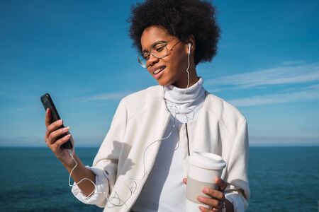 Portrait of young beautiful latin woman using her mobile phone with earphones and holding a cup of...
