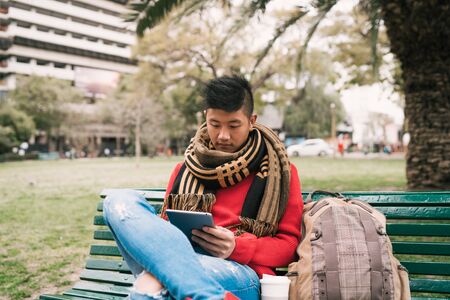 Portrait of young Asian man using his digital tablet while sitting in a bench outdoors. Technology...