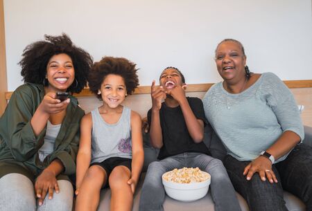 Portrait of African American grandmother, mother and children watching a movie and eating popcorn while sitting on sofa at home. Family and lifestyle concept.の写真素材
