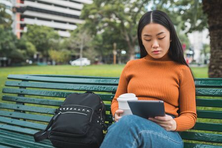 Portrait of young Asian woman using her digital tablet while holding a cup of coffee in the park outdoors.の写真素材