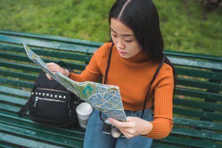 Portrait of young Asian woman sitting on bench in the park and looking at a map. Travel concept....