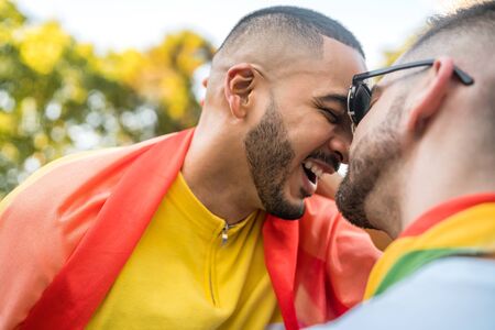 Portrait of young gay couple embracing and showing their love with rainbow flag in the stret. LGBT and love concept.の写真素材