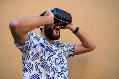 Portrait of young man playing with VR-headset glasses of virtual reality against yellow background....