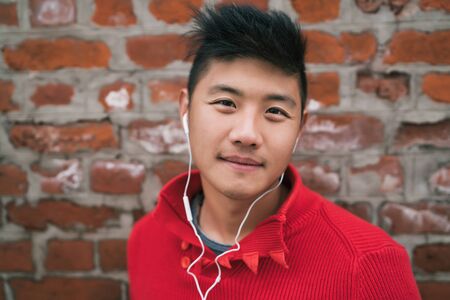 Portrait of young Asian boy listening to music with earphones outdoors against brick wall. Urban concept.の写真素材